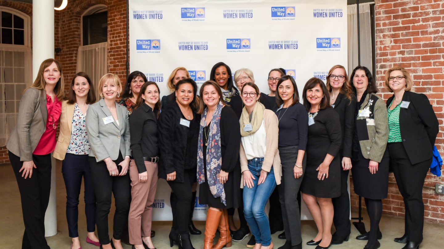 A group of smiling Women United members standing in front of a step-and-repeat featuring Women United and United Way of Rhode Island logos.