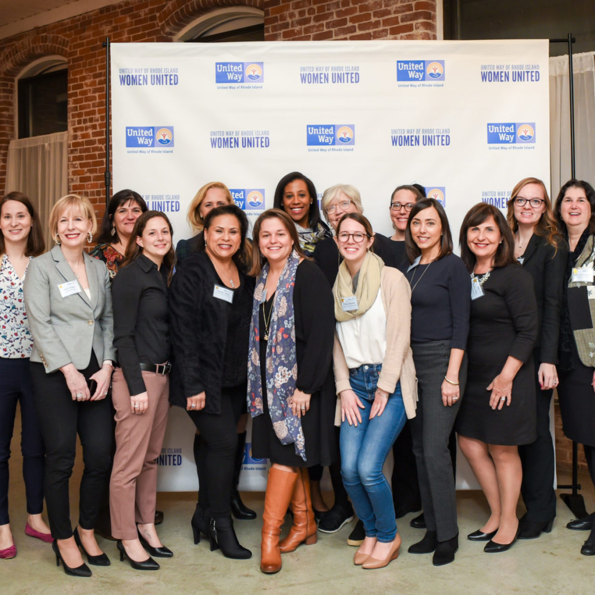A group of smiling Women United members standing in front of a step-and-repeat featuring Women United and United Way of Rhode Island logos.