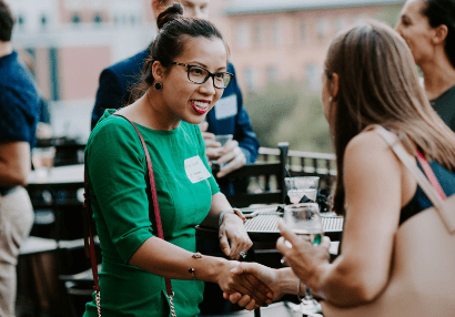 Two women shaking hands during a previous Young Leaders Circle networking event, while others mingle in the background.