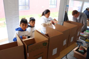 Two children pose behind boxes of books. An adult looks at a book next to them.