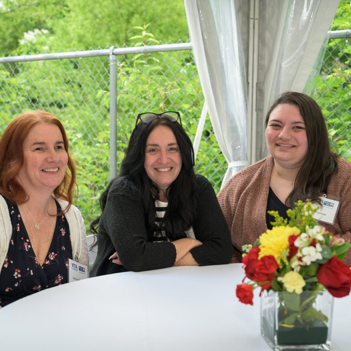 Three smiling attendees sit at a round table in front of trees.
