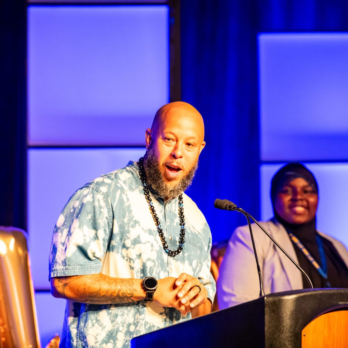 Host Chachi Carvalho speaking from a podium with co-host Wujuudat Balogun smiling in the background.