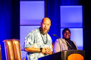 Host Chachi Carvalho speaking from a podium with co-host Wujuudat Balogun smiling in the background.