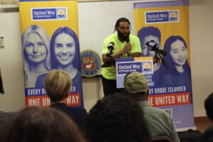 A Black man wearing a neon yellow T-shirt and speaking from a podium.